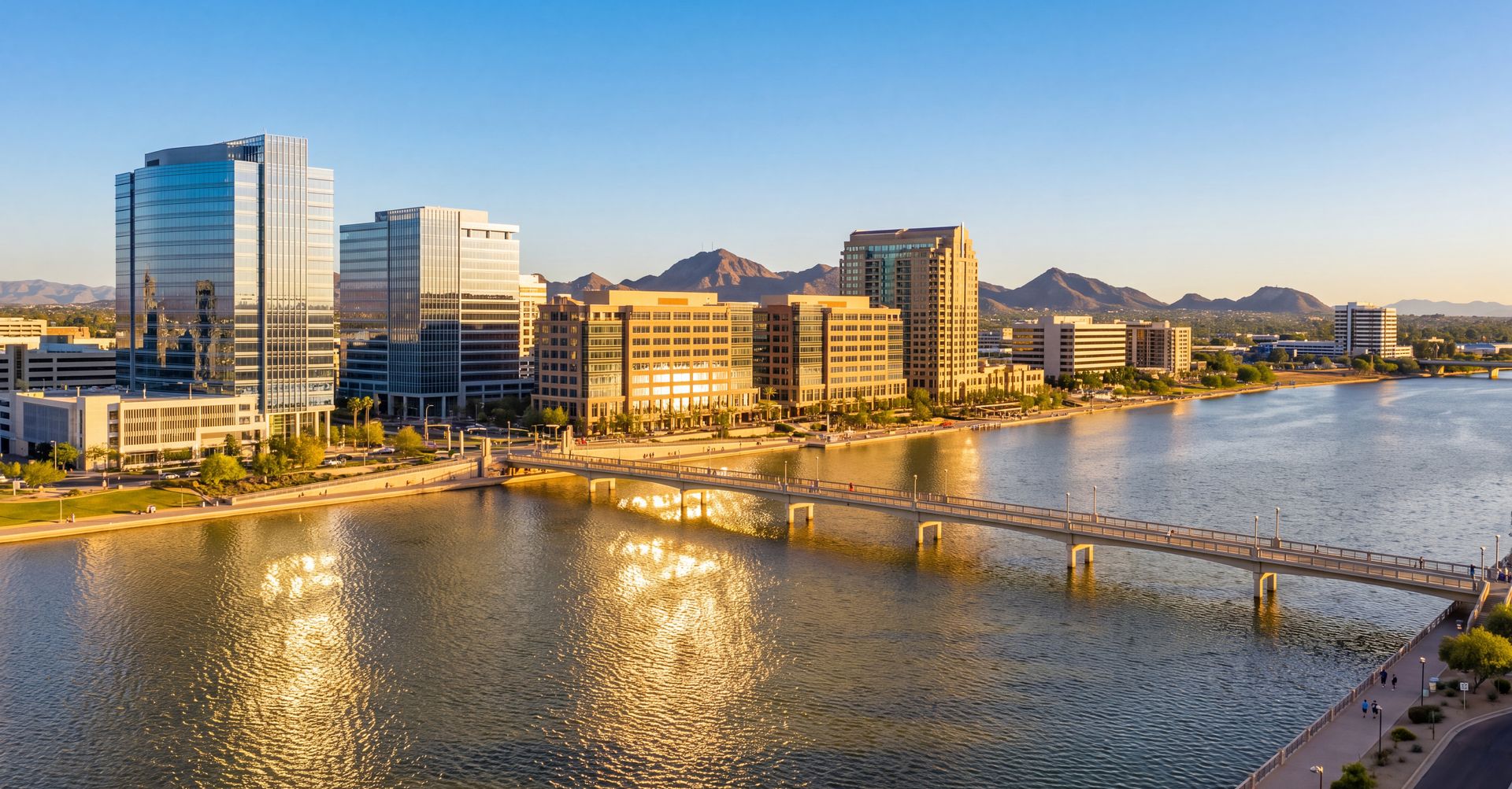 Glass offices along Tempe Town Lake with the lake's pedestrian bridge and desert mountains under a clear Arizona sky